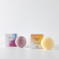 MATER Beauty HAIR Ritual set, T.L.C. Nourishing Shampoo Bar and GOOD HAIR DAYS, Supercharged Conditioner Bar. Two round pucks, pale purple and yellow, next to the cardboard boxes they come in.